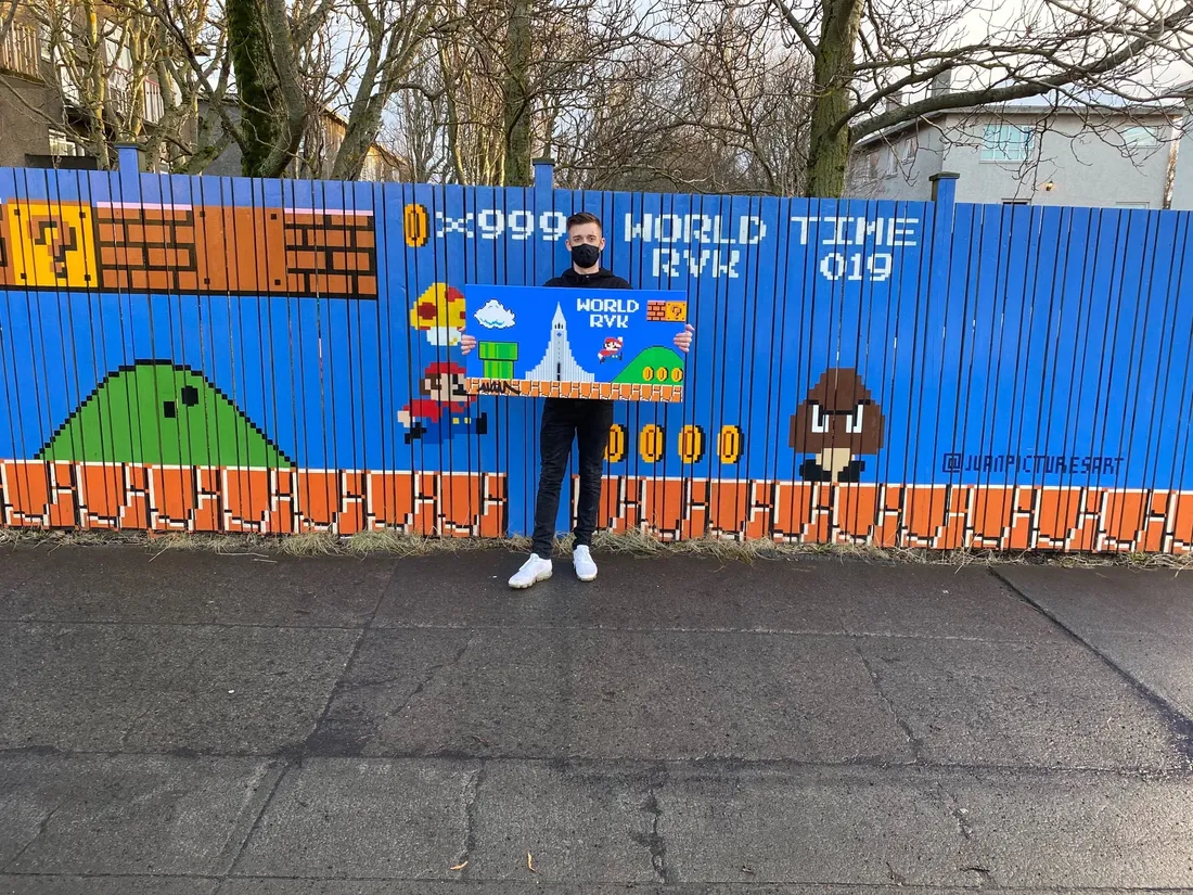 Juan standing in front of his World RVK mural — a Super Mario level painted across a blue fence — holding a framed print of the same scene.